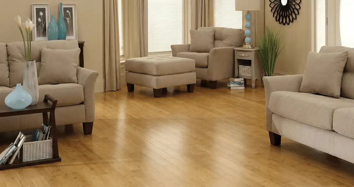 A living room with professionally installed bamboo flooring, beige sofas, a matching ottoman, a side table with a blue lamp, vases, framed pictures, and large windows with blinds and curtains in El Paso, TX